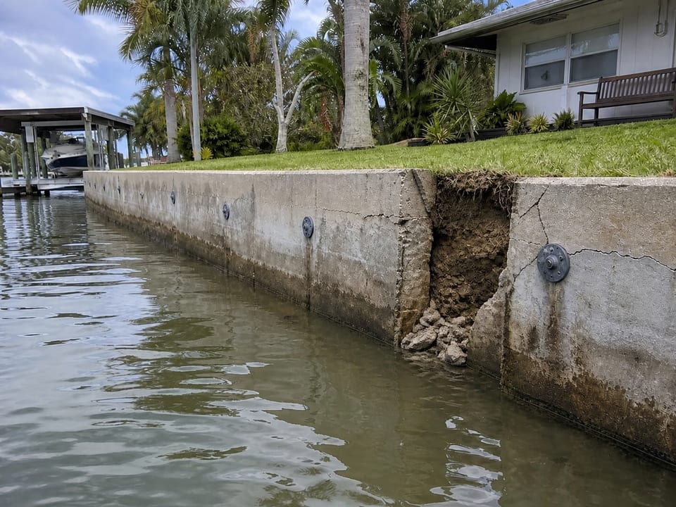 Illustration of a cracked concrete seawall with an irregular break and exposed soil beside a waterfront home.