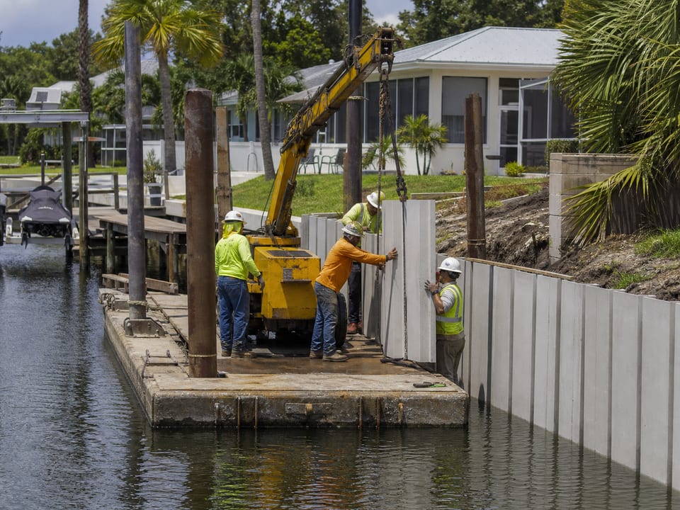 Illustration of workers installing vertical seawall panels along a residential canal.